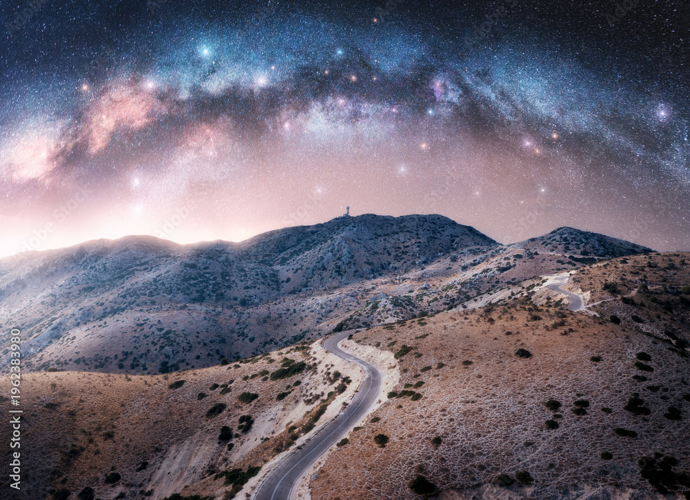 Fototapeta premium Milky Way arch and winding mountain road at night. Sky with arched milky way and bright stars in summer. Lefkada, Greece. Top view of empty road, hills. Beautiful landscape with highway, space. Travel