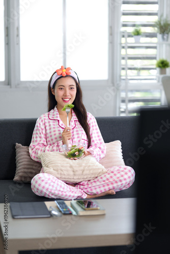 Asian young woman healthy eating salad cozy pajamas relaxing home living room enjoying wholesome breakfast wellness lifestyle at apartment