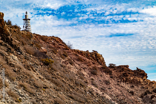 Rocky slope with arid soil and wild vegetation with a lighthouse at top, blue sky with abundant clouds on Coronado Island in the Loreto Bay National Marine Park in Baja California Sur, Mexico