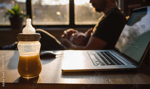 Warm baby bottle with formula milk propped next to a working laptop, and a young father caring and feeding his infant in background. Modern fatherhood concept as balance of parenting and remote work