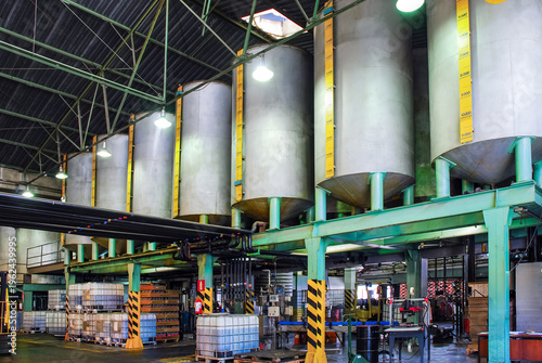 Industrial facility featuring large on a green metal structure, with IBC totes, barrels, and equipment below in a warehouse setting. Focused on chemical or oil processing.