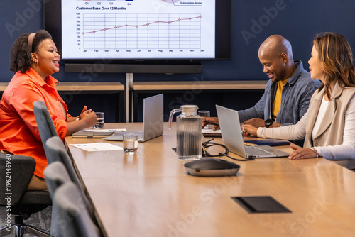 Diverse team meeting around long table in boardroom in business casual with laptops, speaker, chart