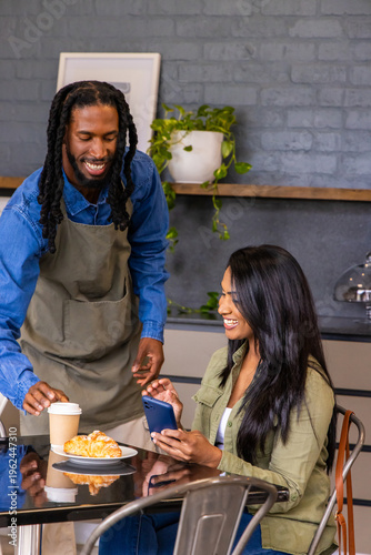 African American barista in denim apron handing coffee cup to Indian customer with phone at cafe