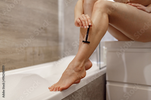 Beautiful young woman shaving legs with razor blade in bathroom while sitting on the edge of the sink, close up. Beauty concept