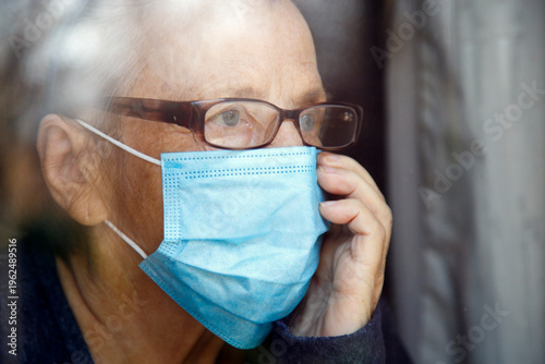 Senior woman wearing face mask looking out of glass window during covid - 1 9 pandemic. Portrait of an elderly woman in quarantine wearing a mask.