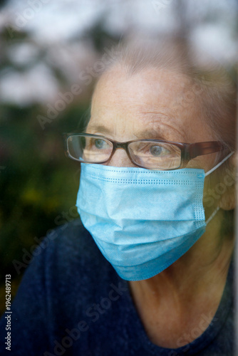 Senior woman wearing face mask looking out of glass window during covid - 1 9 pandemic. Portrait of an elderly woman in quarantine wearing a mask.