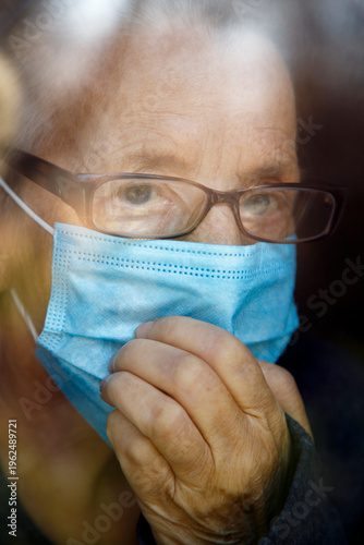 Senior woman wearing face mask looking out of glass window during covid - 1 9 pandemic. Portrait of an elderly woman in quarantine wearing a mask.