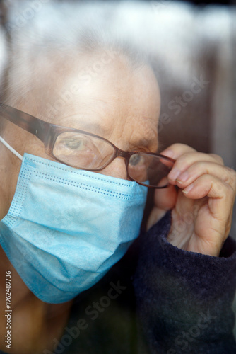 Senior woman wearing face mask looking out of glass window during covid - 1 9 pandemic. Portrait of an elderly woman in quarantine wearing a mask.