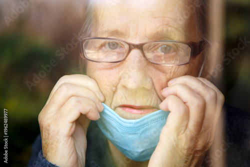 Portrait of an elderly woman in a window after quarantine with her mask removed. An old woman in a window with her mask down, after the pandemic.