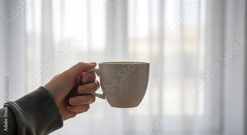 Close up of a person holding a white mug full of hot beverage in front of sheer curtains