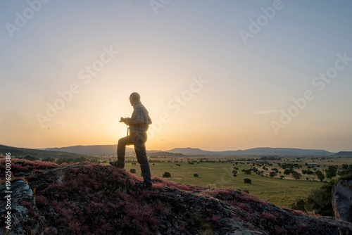 Billede på lærred silhouette of a man photographing in the mountains, A person is silhouetted against the setting sun while walking along a rocky, flower-covered ridge overlooking a vast landscape