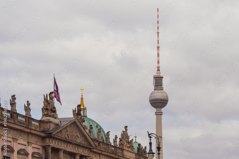 Obraz premium Berlin TV Tower stands tall against cloudy sky. Historic building with ornate roof and flag flies beside tower. No people visible. Urban skyline image ideal for travel, architecture