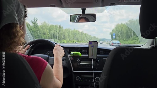 Woman is driving a car with a green device holder on the dashboard. The device is a phone charger