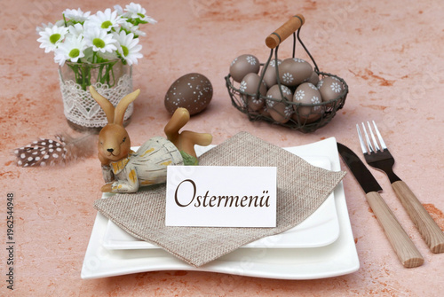 Harmonische Tischdekoration zu Ostern. Tischdekoration mit Osterhase,  Karte mit dem Schrifzug Ostermenü auf einem Teller, Besteck und weiße Gänseblümchen.