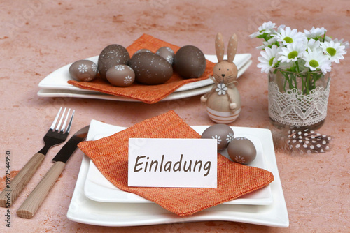 Tischgedeck mit weißen Tellern, orangefarbenen Servietten, braunen Ostereiern, einem kleinen Hasen aus Holz mit weißen Blumen und dem Schriftzug Einladung auf einer Tischkarte.