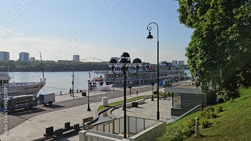 MOSCOW - JUL 26 , 2025: City street with a river running through it. The street is lined with trees and has a few benches. A boat is docked in the river Stalin era riverside station