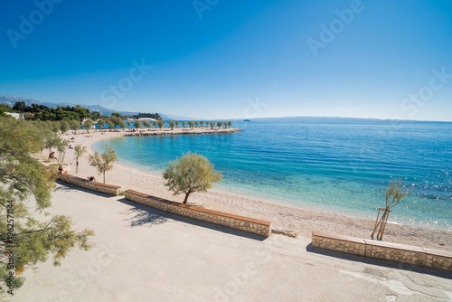 Wide-angle view of Znjan beach in Split, Croatia. Scenic coastline with stone benches, green trees, and crystal clear blue Adriatic sea under a bright sunny sky.