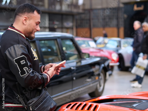 Billede på lærred Profile of a bearded man in a black leather varsity jacket taking a photo
with a pink smartphone
