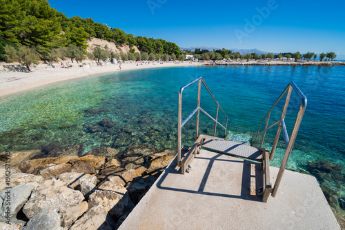 Metal stairs leading into the turquoise Adriatic Sea at a pebble beach in Dalmatia, Croatia, near Split. Surrounded by pine trees under a clear blue sky.