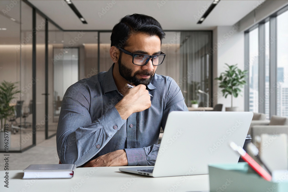 Obraz premium Young serious indian professional business man, focused ethnic male student wearing glasses working on laptop, remote studying using computer looking at screen watching seminar webinar at home office.