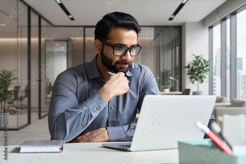 Konstfotografi Young serious indian professional business man, focused ethnic male student wearing glasses working on laptop, remote studying using computer looking at screen watching seminar webinar at home office