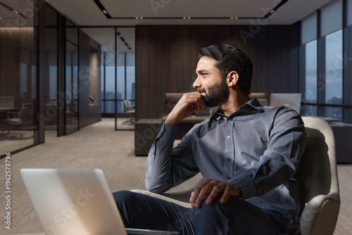 Young thoughtful smart indian professional business man executive looking away relaxing sitting on chair in modern office lobby with laptop, thinking of new ideas, dreaming of success, planning.