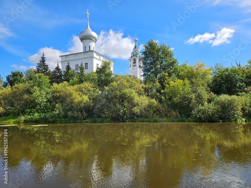 Large white building with a blue dome sits in front of a lush green forest. The water in the foreground is calm and reflective, creating a peaceful atmosphere