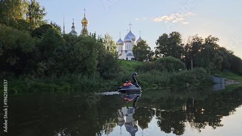 Man is riding a boat on a lake near a church. The sky is blue and the water is calm