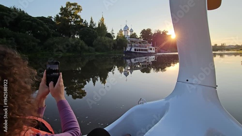 Woman is sitting on a boat and taking a picture of the sunset. The water is calm and the sky is orange and pink