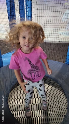 Young girl is standing in a blue and white bounce house trampoline . She is wearing a pink shirt and white pants