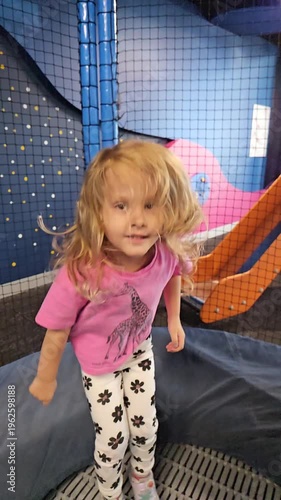 Young girl with long blonde hair is wearing a pink shirt and white pants. She is standing in front of a blue wall bounce trampoline with a giraffe on her shirt