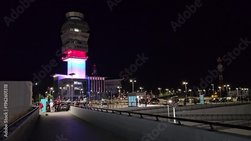 BELGRADE - SEP 18, 2025: City street at night with tall building in the background. The building is lit up in red, blue, white. There are several people walking around, some of them carrying handbags