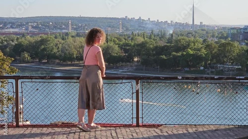 Woman stands on a bridge overlooking a body of water. The scene is peaceful and serene, with the woman taking in the view and enjoying the moment