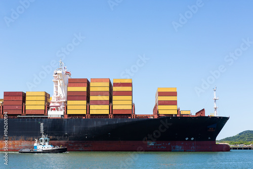 Massive black freighter stacked with vibrant red and yellow containers docked at Santos Port, Brazil. Blue tugboat maneuvers nearby on calm waters under clear blue sky, green hills visible, ideal for 