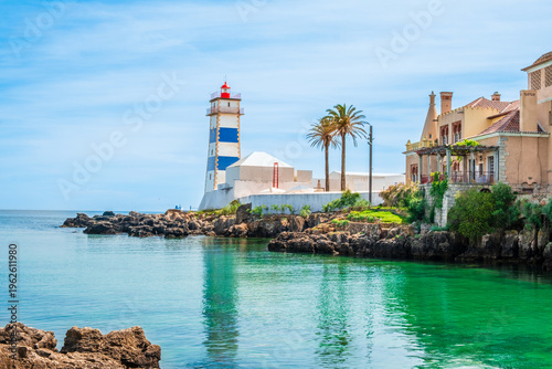 Santa Marta lighthouse and rocky coastline with palm tree in Cascais, Portugal. White and blue lighthouse, clear sea water and historic buildings in bright sunlight