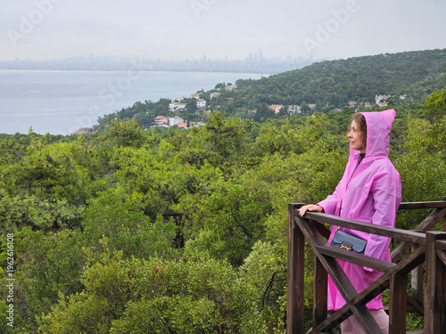 Wallpaper Mural Woman in a pink raincoat stands on a wooden platform overlooking a forest. The sky is cloudy and the water is in the background Torontodigital.ca