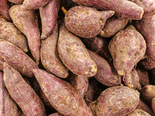 Close-up of fresh purple sweet potatoes stacked together at a market, showing rough natural skin texture. Nutritious root vegetables rich in fiber, antioxidants, and commonly used in tropical cuisine.
