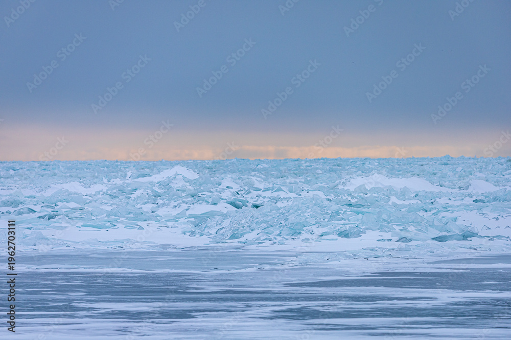 Obraz premium A breathtaking winter landscape featuring vast piles of translucent blue ice shards pushed up onto the shoreline of a frozen Great Lake. In the distance, a soft band of peach-colored light glows.