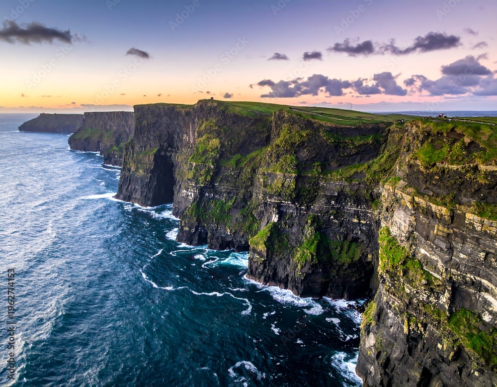 Obraz premium Aerial view of high green cliffs meeting vast blue ocean under a colorful sunset