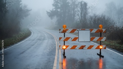 A wet winding road with a construction barrier and warning lights on a foggy day traffic safety detour closed sign orange white stripes caution hazard obstruction roadwork