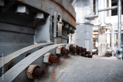 Close-up of rusty bolts on industrial pipe. Heavy machinery in background. Industrial setting with worn metal surfaces. Suitable for engineering, manufacturing, or construction themes
