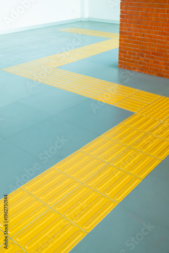Canvas-taulu Interior view showing bright yellow tactile paving guiding path for blind and visually impaired people on a grey tiled floor, adjacent to a textured red brick wall corner