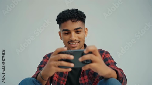 A young man in a red plaid shirt plays a video game on his smartphone.