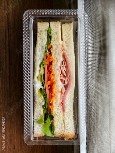 Top view of fresh club sandwich packed with lettuce, carrot, ham, and crab stick in a clear takeaway plastic box on a wooden table. Quick lunch or snack concept