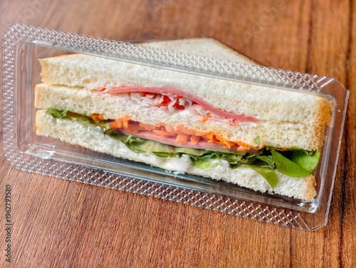 Top view of fresh club sandwich packed with lettuce, carrot, ham, and crab stick in a clear takeaway plastic box on a wooden table. Quick lunch or snack concept