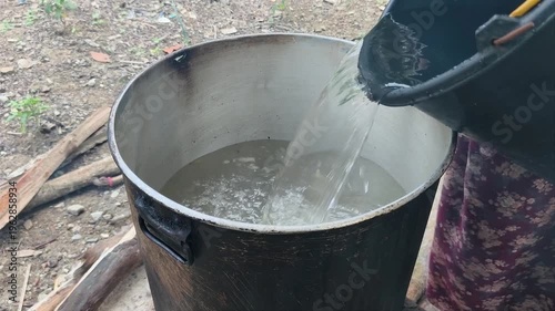 Water is being poured from a dark bucket into a large metal pot.
