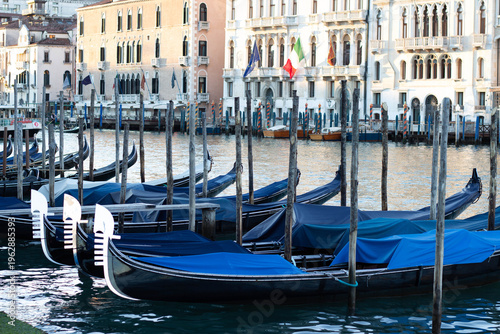 Gondolas with blue covers moored along the canal in Venice, Italy, with historic buildings and flags visible in the background reflecting on the water