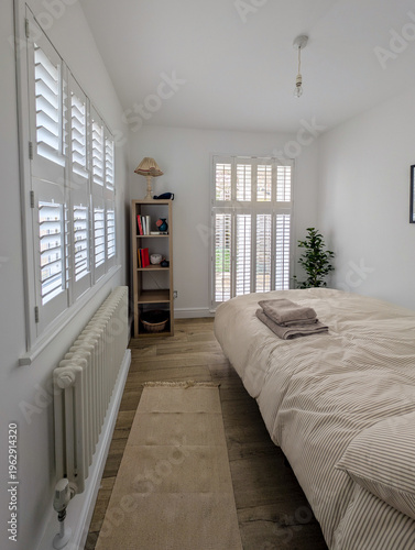 Bright Minimalist Bedroom with Wooden Floor and Natural Light.
A clean and cozy interior featuring a simple bed, soft textiles, indoor plant, and shelving unit. The space is illuminated by daylight 