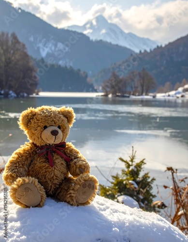 A teddy bear sits on a snowy hill overlooking a serene lake