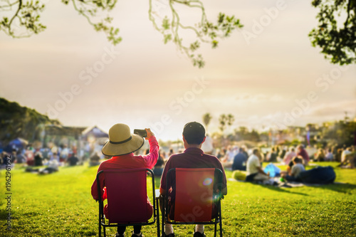 Couple taking photos using smartphone at sunset, unrecognizable crowd of people listen to the music concert in the park.